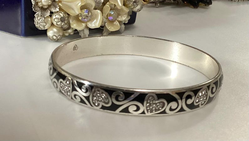 Op de afbeelding: Een zilverkleurige armband met een herhalend patroon van zwarte emaille krullen en hartvormen. De hartjes zijn gevuld met kleine, heldere, fonkelende steentjes. De binnenkant van de metalen band toont een klein gegraveerd keurmerk.