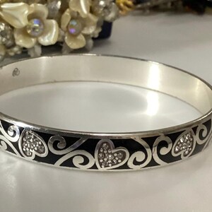Op de afbeelding: Een zilverkleurige armband met een herhalend patroon van zwarte emaille krullen en hartvormen. De hartjes zijn gevuld met kleine, heldere, fonkelende kristallen. De binnenkant van de metalen band is glad en gepolijst met een zilveren afwerking.