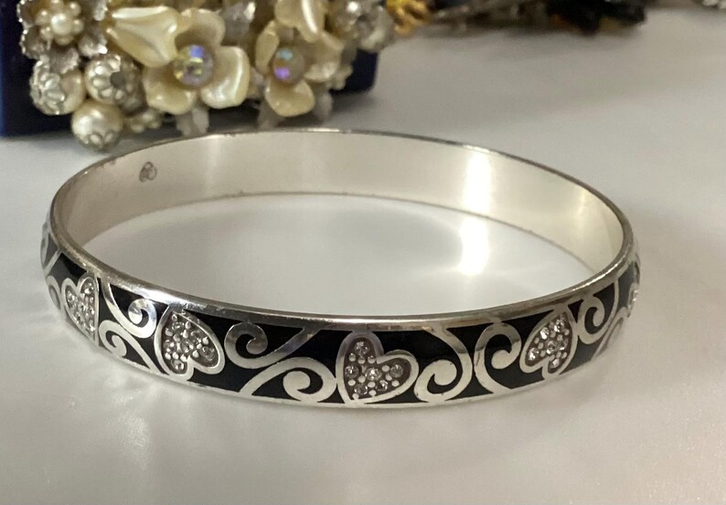Op de afbeelding: Een zilverkleurige armband met een herhalend patroon van zwarte emaille krullen en hartvormen. De hartjes zijn gevuld met kleine, heldere, fonkelende kristallen. De binnenkant van de metalen band is glad en gepolijst met een zilveren afwerking.