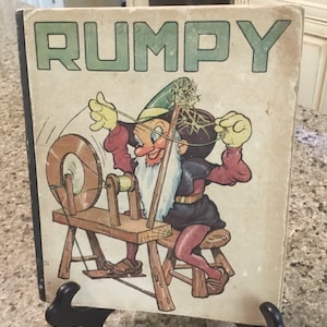 May include: Vintage book titled "RUMPY" with a cartoon illustration of a gnome-like character operating a spinning wheel. The cover is aged with visible wear and tear, and the title is in green block letters.