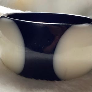May include: A wide, glossy bracelet with a bold design. The bracelet features a striking contrast of black and off-white sections. The smooth, rounded shape and polished finish give it a modern, stylish look. This accessory is perfect for adding a statement to any outfit.