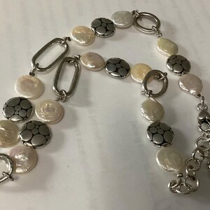 May include: A bracelet featuring alternating white coin-shaped pearls and circular silver-toned metal discs with a dotted pattern. The piece includes elongated oval silver-toned links, a circular toggle clasp, and a short chain extension with a small charm.