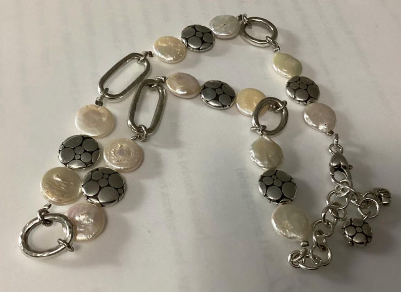May include: A bracelet featuring alternating white coin-shaped pearls and circular silver-toned metal discs with a dotted pattern. The piece includes elongated oval silver-toned links, a circular toggle clasp, and a short chain extension with a small charm.