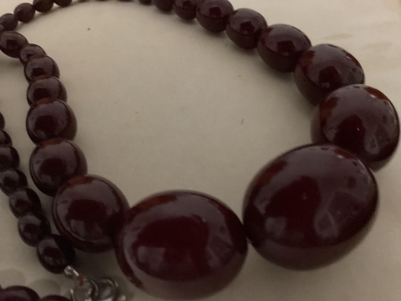Op de afbeelding: Een ketting met gegradueerde, gepolijste, diepbordeauxrode kralen. De kralen worden groter naar het midden van de streng toe, die is vastgezet met een zilverkleurige metalen sluiting die zichtbaar is aan het uiteinde van de ketting.