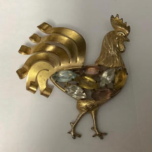 Op de afbeelding: Een goudkleurige metalen haanbroche met een gestileerde staart van gebogen linten en een lichaam versierd met marquise-geslepen strass-steentjes in lichtblauw, helder, geel en zachtroze, met een getextureerde kam en gedetailleerde poten.