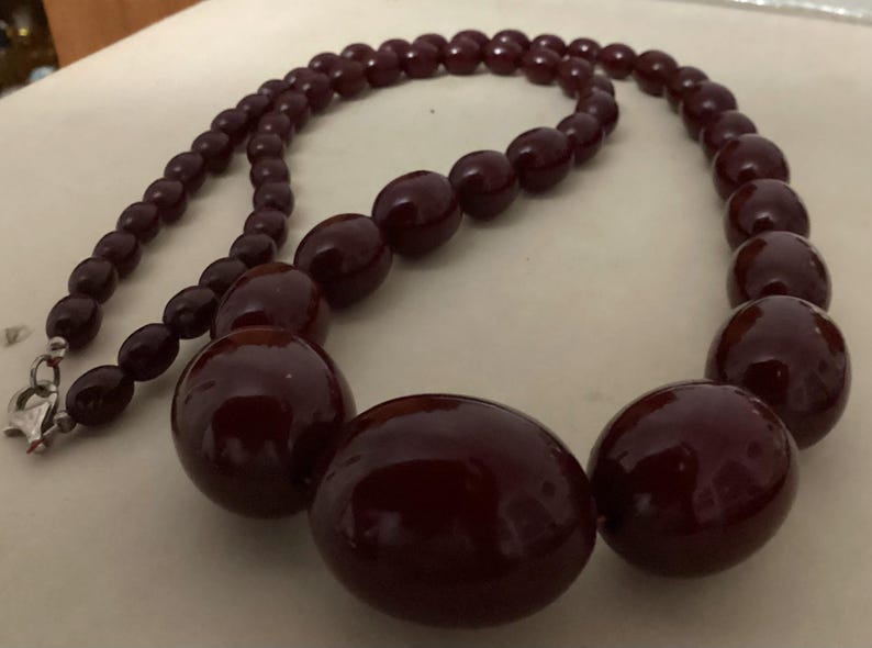 Op de afbeelding: Een ketting met gegradueerde, gepolijste, diep bordeauxrode kralen. De ketting verloopt van kleinere, uniforme kralen aan de uiteinden naar aanzienlijk grotere, ronde kralen in het midden, vastgezet met een zilverkleurige metalen sluiting.