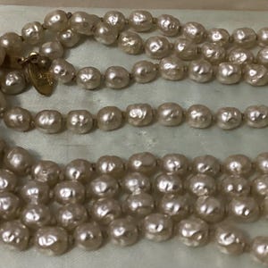 May include: A vintage pearl necklace with three strands of irregular, off-white pearls. The necklace is displayed in a light blue box, showcasing the classic design and texture of the pearls. The clasp is gold-toned.