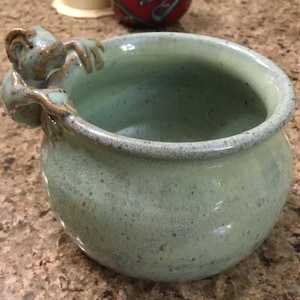Puede incluir: Un cuenco de cerámica hecho a mano en tonos verdes y marrones. El cuenco presenta una figura de mono esculpida agarrada al borde. El cuenco tiene una textura moteada y una forma redondeada, adecuada para servir o exhibir.