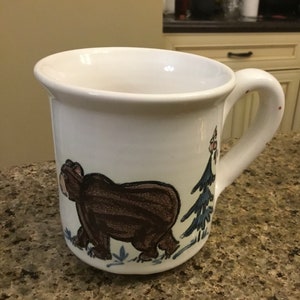 Puede incluir: Taza de cerámica blanca con una ilustración de oso pardo y un diseño de árbol azul. La taza tiene un asa curva y un borde ancho. El oso está representado caminando, y el árbol es una conífera estilizada.