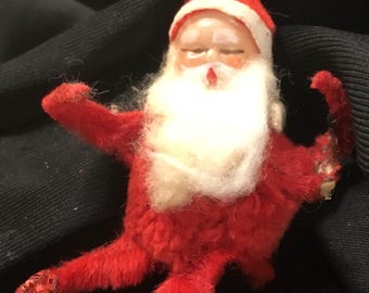 May include: A vintage Christmas ornament in the shape of Santa Claus. The ornament is red with a white beard and hat trim. The face is painted with closed eyes and a rosy nose. A gold string is attached for hanging.