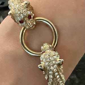 May include: A gold-tone metal bracelet featuring two stylized feline heads encrusted with clear rhinestones and small red stones for eyes. The two heads are connected by a smooth, circular gold-tone ring, creating a continuous, hinged design.