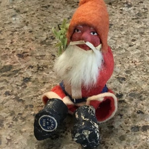 May include: A vintage Christmas gnome doll with a red felt hat and coat, white beard, and black boots. The doll has a painted face and is holding a small green sprig. The boots are marked "Made in Germany."