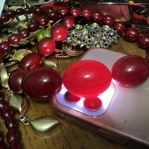 Op de afbeelding: Een collectie sieraden met een opvallende streng dieprode, gepolijste, ovale kralen. De kralen zijn gerangschikt naast een smartphone met een oplichtende cameraflitser, een zilverkleurige broche met groene accenten en goudkleurige metalen bladhangers.