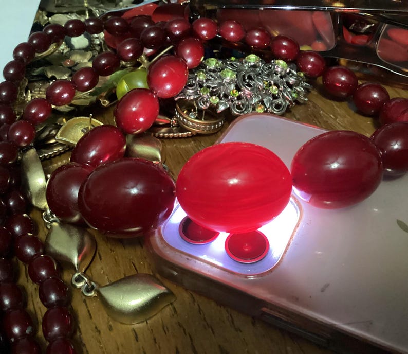 Op de afbeelding: Een collectie sieraden met een opvallende streng dieprode, gepolijste, ovale kralen. De kralen zijn gerangschikt naast een smartphone met een oplichtende cameraflitser, een zilverkleurige broche met groene accenten en goudkleurige metalen bladhangers.