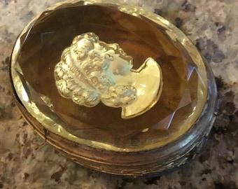 May include: An oval antique jewellery box with a gold-coloured metal frame and a faceted, clear top. The box features a gold-coloured relief of a classical profile. The interior is a warm brown colour.