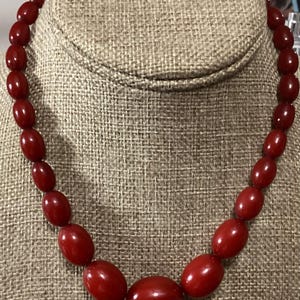 May include: A single-strand necklace featuring smooth, polished, deep red oval beads. The beads are graduated in size, with the largest bead positioned at the center and smaller beads extending symmetrically toward the back of the necklace.