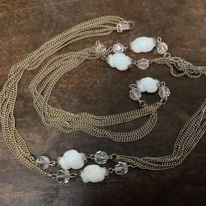 May include: A gold-coloured necklace with multiple strands of delicate chain. The necklace features white, cloud-shaped beads and clear, round beads. The clasp is visible at the top. The jewellery is displayed on a dark brown surface.