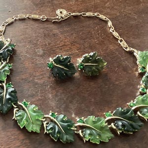 May include: A vintage jewelry set featuring a necklace and matching earrings. The necklace and earrings are designed with green leaf-shaped pendants and gold-tone metal accents. Each leaf is adorned with small green gemstones.
