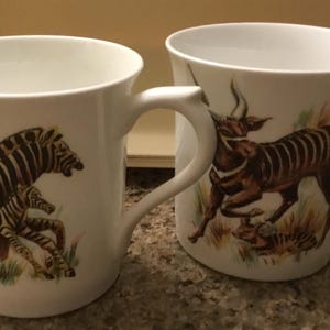 May include: Two white ceramic mugs with animal illustrations. One mug features a zebra and a baby zebra, the other depicts a brown antelope. Both mugs have curved handles and are set against a neutral background.