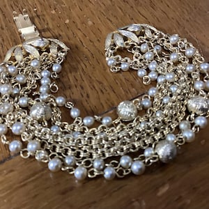 Könnte beinhalten: Ein mehrreihiges goldfarbenes Armband mit zahlreichen kleinen weißen Kunstperlen, die mit strukturierten goldfarbenen Metallperlen durchsetzt sind. Das Armband besteht aus mehreren feinen Gliederketten und hat einen blattförmigen Verschluss.
