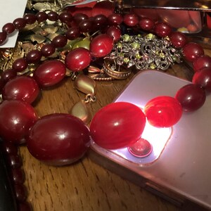 Op de afbeelding: Een ketting bestaande uit gegradueerde, dieprode, gepolijste, ovaalvormige kralen. De kralen vari&euml;ren in grootte, met grotere, afgeronde stukken in het midden. De ketting ligt over een lichtgekleurde smartphone, die een fel wit licht uitstraalt vanuit de cameralens, waardoor het doorschijnende rode oppervlak van de kralen wordt verlicht.