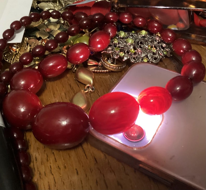 Op de afbeelding: Een ketting bestaande uit gegradueerde, dieprode, gepolijste, ovaalvormige kralen. De kralen vari&euml;ren in grootte, met grotere, afgeronde stukken in het midden. De ketting ligt over een lichtgekleurde smartphone, die een fel wit licht uitstraalt vanuit de cameralens, waardoor het doorschijnende rode oppervlak van de kralen wordt verlicht.