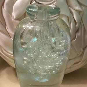 May include: A clear glass perfume bottle with a rounded body and a bulbous stopper. The bottle is filled with a light-colored liquid and iridescent bubbles. The background is a soft, white fabric.