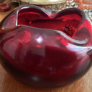 May include: A deep red, heart-shaped glass bowl. The bowl has a smooth, glossy surface and a rounded shape. The interior of the bowl is also red, and it appears to be filled with a liquid or gel. A small, red, round object is inside the bowl.