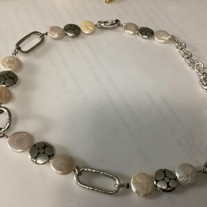 May include: A necklace featuring a sequence of round, iridescent white coin-shaped beads alternating with textured silver-toned metal discs and elongated oval silver-toned links. The piece includes a silver-toned chain extender and a lobster clasp closure.