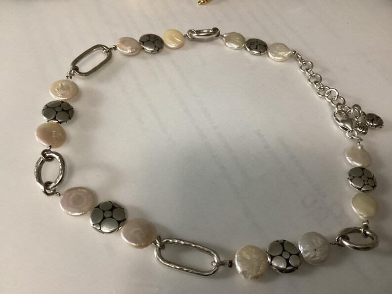 May include: A necklace featuring a sequence of round, iridescent white coin-shaped beads alternating with textured silver-toned metal discs and elongated oval silver-toned links. The piece includes a silver-toned chain extender and a lobster clasp closure.