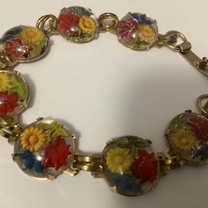 May include: A vintage gold-tone bracelet featuring clear, round resin charms with colorful floral arrangements. Each charm showcases red, yellow, and blue flowers. The bracelet has a clasp closure.