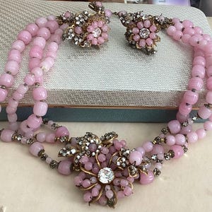 Puede incluir: Un collar de cuentas rosa vintage y un juego de pendientes a juego. El collar presenta tres hilos de cuentas rosas con un colgante floral y detalles de pedrería. Los pendientes tienen un diseño floral similar. Un conjunto de joyas clásico.