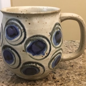 Może przedstawiać: Ręcznie wykonany ceramiczny kubek z nakrapianym, kremowym szkliwem. Kubek posiada serię niebieskich i szarych okrągłych wzorów. Uchwyt ma solidny, zaokrąglony kształt. Ten wyjątkowy kubek jest idealny do kawy lub herbaty.