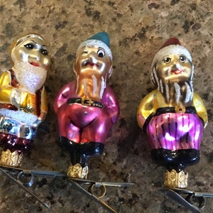 Könnte beinhalten: Drei Vintage-Weihnachtsschmuck in Form von Zwergen. Jeder Zwerg hat ein andersfarbiges Outfit, darunter Rot, Rosa und Lila. Sie haben weiße Bärte und Hüte und sind an Metallclips zum Aufhängen befestigt.