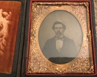 May include: Vintage portrait of a man in formal attire, including a bow tie, set within an elaborate gold-toned frame. The photograph is enclosed in a black case with a velvet lining, representing historical photography.