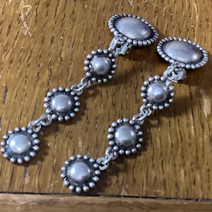 May include: A pair of silver-toned clip-on earrings featuring a vertical drop design. Each earring consists of four circular, bead-edged components connected by small metal links, with a larger oval-shaped top piece securing the clip mechanism.