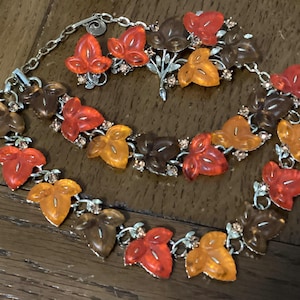 May include: A vintage jewelry set featuring a necklace, bracelet, and earrings. The set showcases leaf-shaped pendants in shades of orange, red, and brown, accented with small, sparkling rhinestones. The metal components have a silver finish.