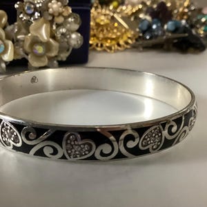 Op de afbeelding: Een zilverkleurige armband met een herhalend patroon van zwarte emaille harten en krullende filigraan details. De metalen band heeft een gladde, gepolijste afwerking en een ronde vorm, met een consistent ontwerp rondom de gehele omtrek.