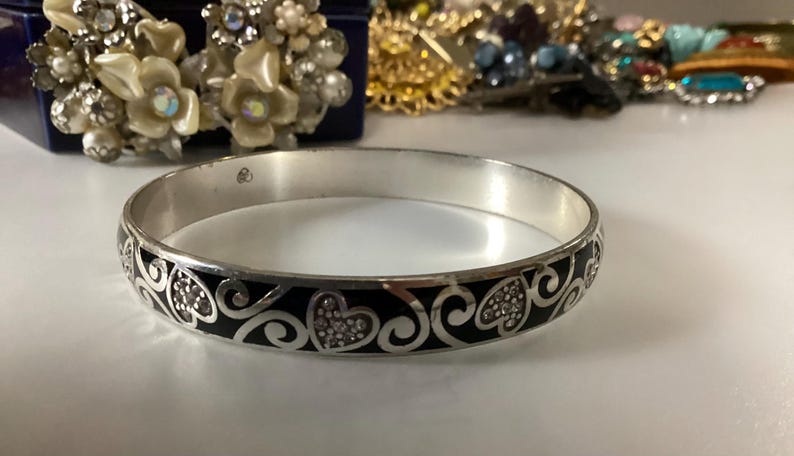 Op de afbeelding: Een zilverkleurige armband met een herhalend patroon van zwarte emaille harten en krullende filigraan details. De metalen band heeft een gladde, gepolijste afwerking en een ronde vorm, met een consistent ontwerp rondom de gehele omtrek.