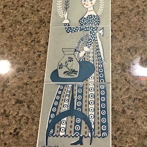 May include: A set of three ceramic tiles featuring a stylized illustration of a woman in a long dress holding a flower and a vase. The design uses blue, white, and brown on a gray background. The tiles are arranged vertically.