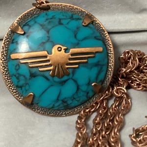 Pode incluir: Colar com pingente em tom de cobre com uma pedra turquesa redonda e um design Thunderbird em cobre. O pingente tem uma borda de cobre texturizada e uma corrente de cobre combinando.