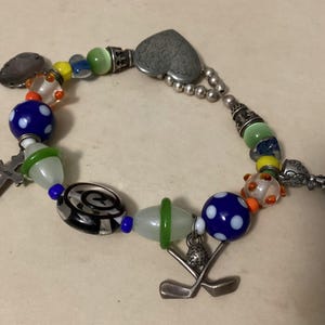May include: A beaded bracelet with a variety of charms and beads. The bracelet includes a silver heart-shaped charm, a cross, and golf-themed charms. The beads are colorful, including blue polka-dot beads, and various glass beads.