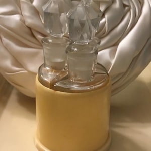 Peut inclure: Un flacon de parfum vintage avec deux bouchons en verre transparent avec des dessus facettés. Le flacon a une base jaune et un dessus en verre transparent. Le fond est un tissu crème.