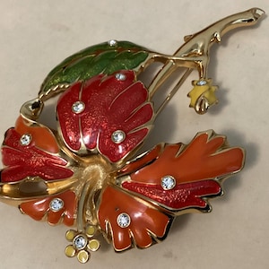 May include: A gold-toned floral brooch with red, orange, and green enamel detailing. The brooch features a flower and leaf design, accented with clear rhinestones. The brooch is a vintage style.
