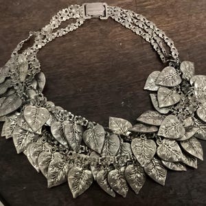 May include: Silver-toned necklace featuring a leaf design. The necklace has multiple strands and a clasp closure. The leaves are detailed with veining and hang from the chain. The jewelry is displayed on a dark wooden surface.