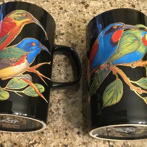 May include: Two black ceramic mugs with colorful bird illustrations. One mug features red, orange, and blue birds, while the other showcases blue, green, and red birds. Both mugs have black handles and a gold-colored trim.