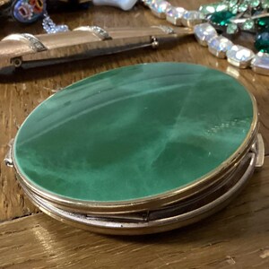 May include: An oval compact mirror featuring a smooth, solid green top surface with subtle marbled variations. The compact has a metallic gold-toned rim and a hinged construction, presenting a sleek, rounded profile with a polished finish.