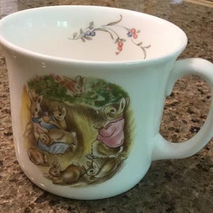 May include: White ceramic mug featuring a charming illustration of rabbits in a garden setting. The mug has a curved handle and a floral design near the rim. The artwork depicts rabbits in various poses, with soft colors.