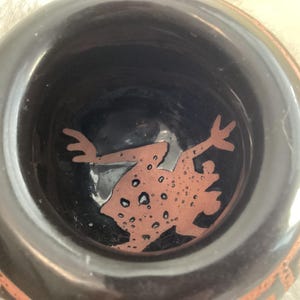 Könnte beinhalten: Eine schwarze Keramikschale mit einem Froschdesign im Inneren. Der Frosch ist braun mit dunkleren Flecken. Der Rand der Schale hat ein dekoratives orangefarbenes Muster. Die Schale wird wahrscheinlich zu dekorativen Zwecken verwendet.