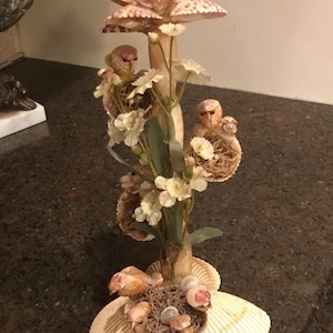 Puede incluir: Una escultura decorativa de conchas marinas con una disposición floral en la parte superior, con pequeñas figuras de pájaros y flores blancas. La base está hecha de conchas grandes. La escultura mide unos 30 cm de altura.